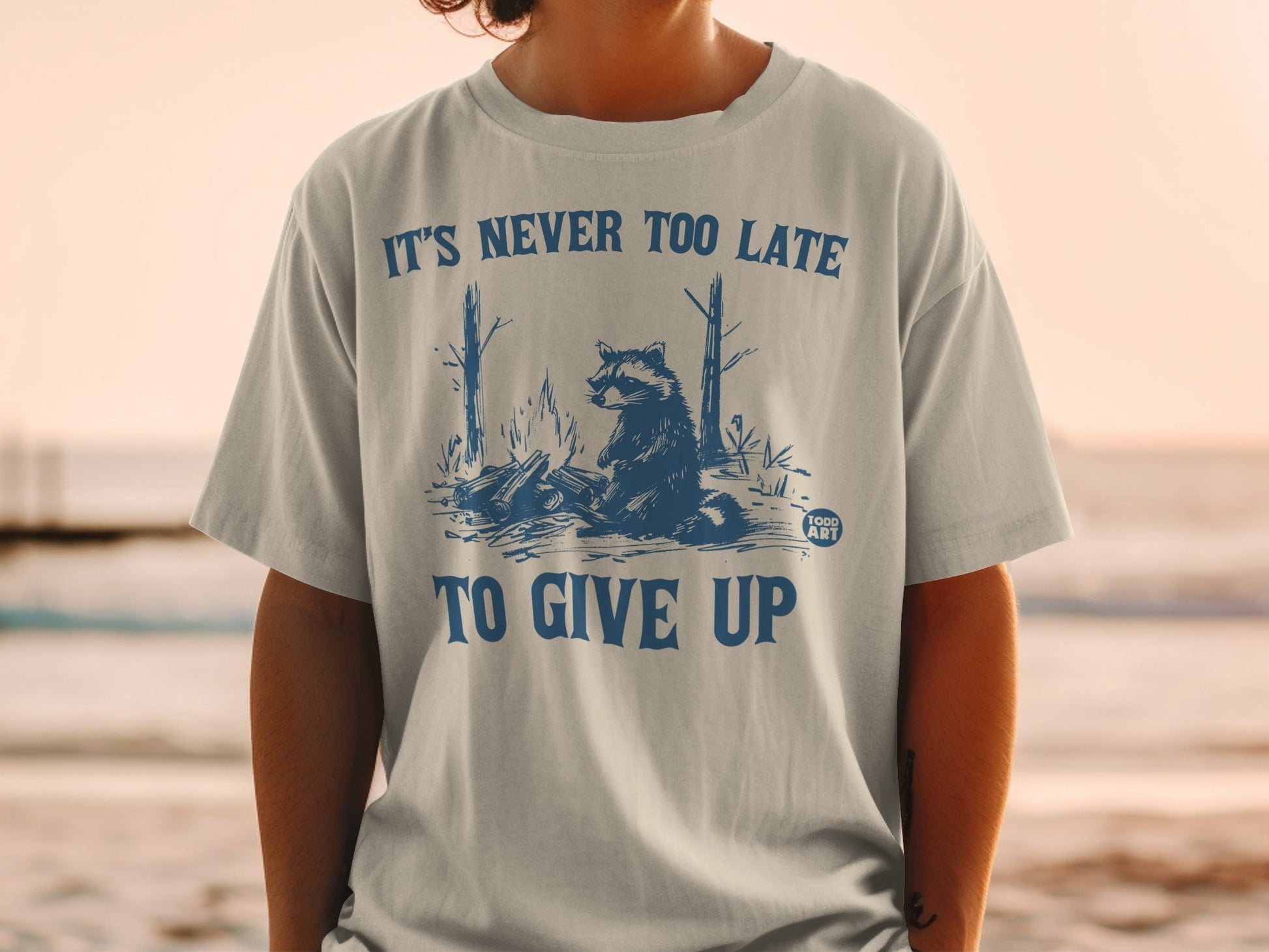 T-shirt with raccoon near campfire and text "IT'S NEVER TOO LATE TO GIVE UP".