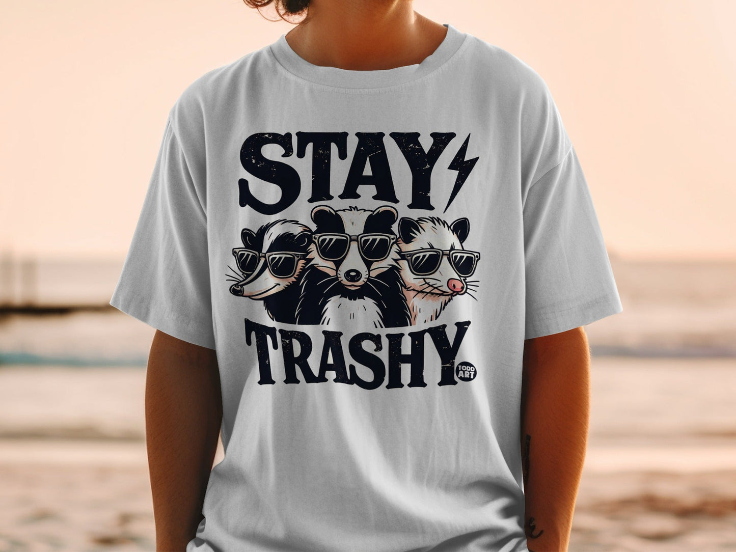 Three cartoon raccoons wearing sunglasses on a white t-shirt with "STAY TRASHY" text.