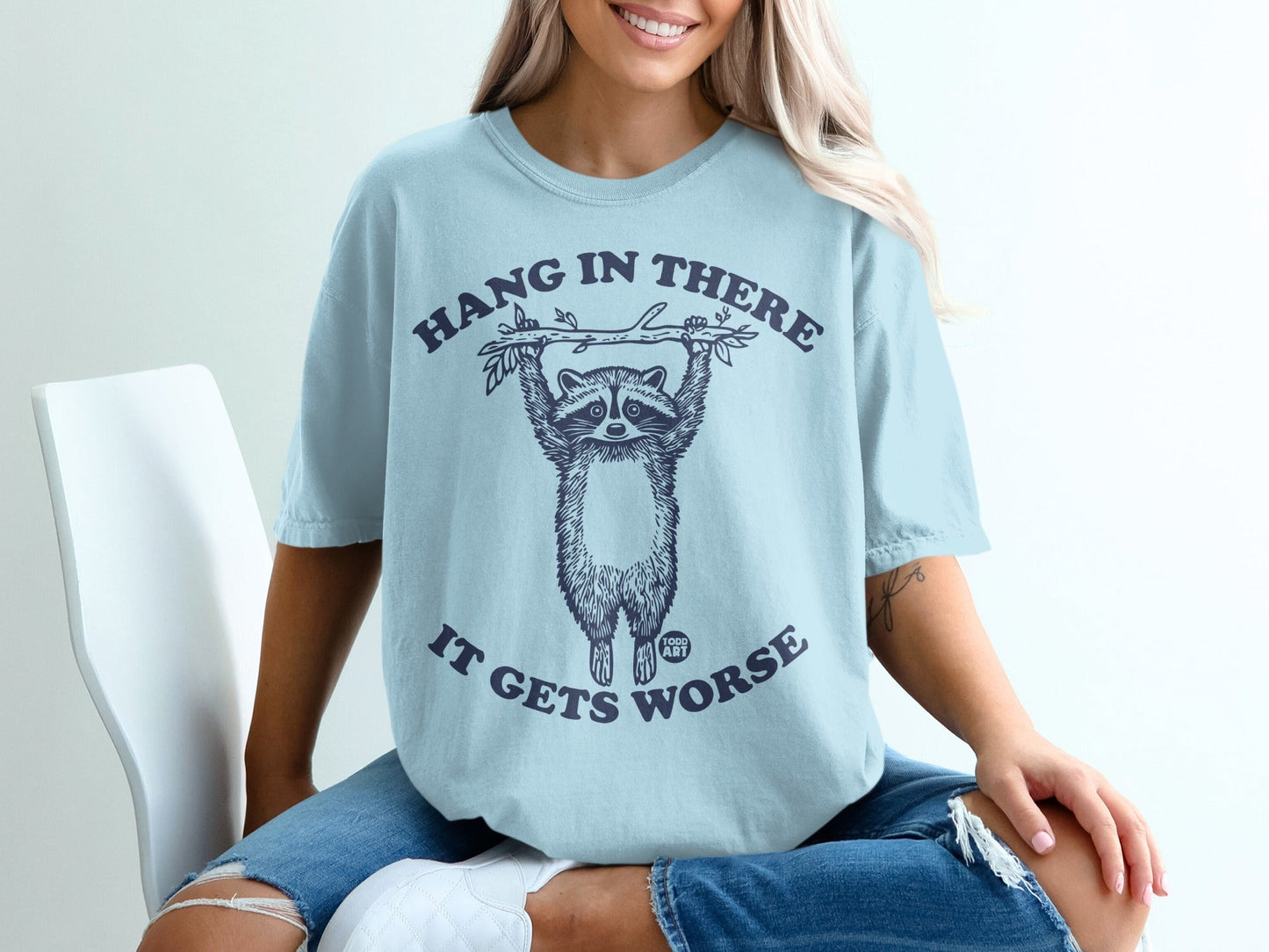 Light blue t-shirt with "Hang in there it gets worse" and a raccoon graphic.