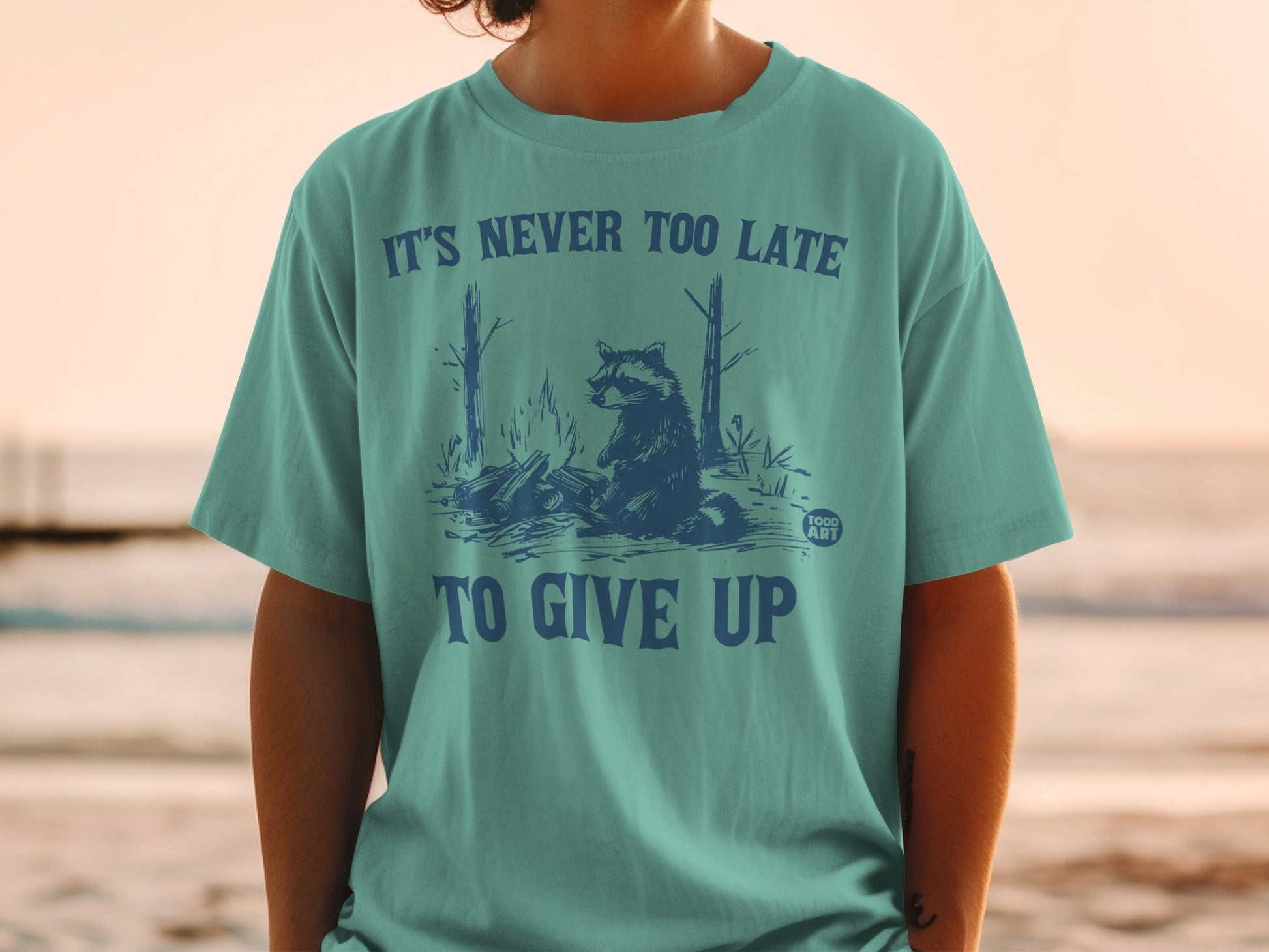 Teal t-shirt with raccoon by campfire graphic and "It's never too late to give up" text.