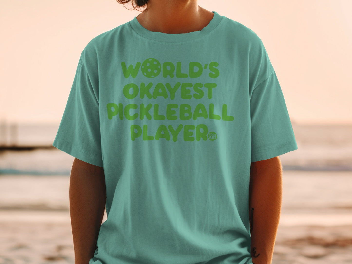 Green t-shirt with "World's Okayest Pickleball Player" text and pickleball graphic.