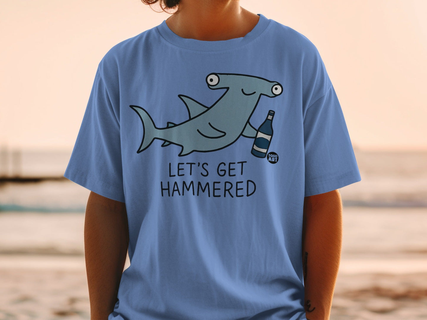 Blue t-shirt with a cartoon hammerhead shark holding a bottle labeled "Food Art".