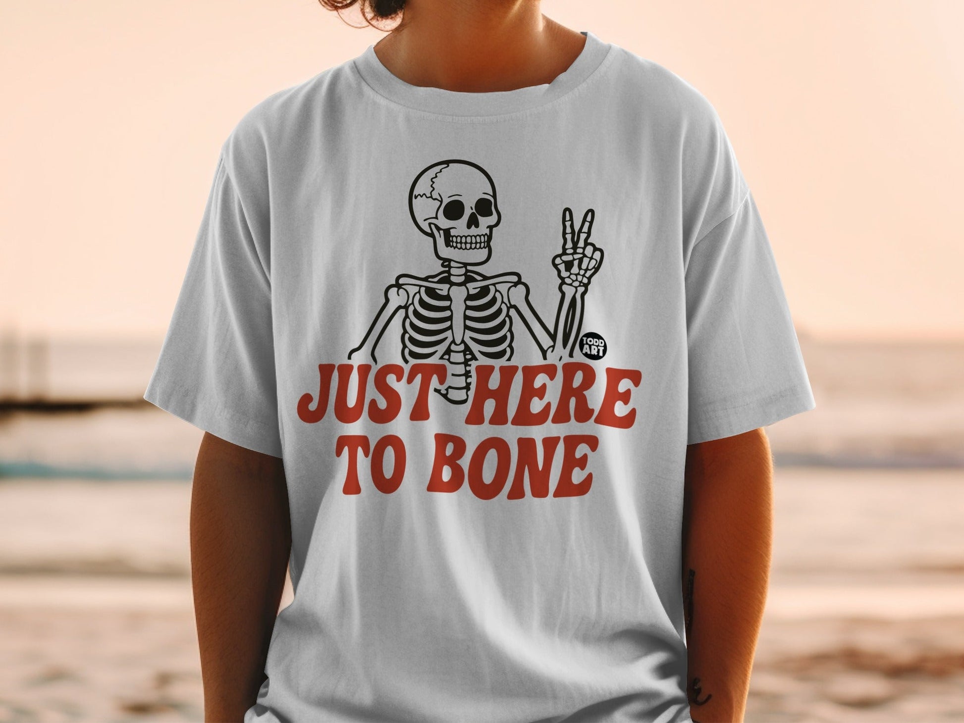 White t-shirt with skeleton making peace sign and text "JUST HERE TO BONE".