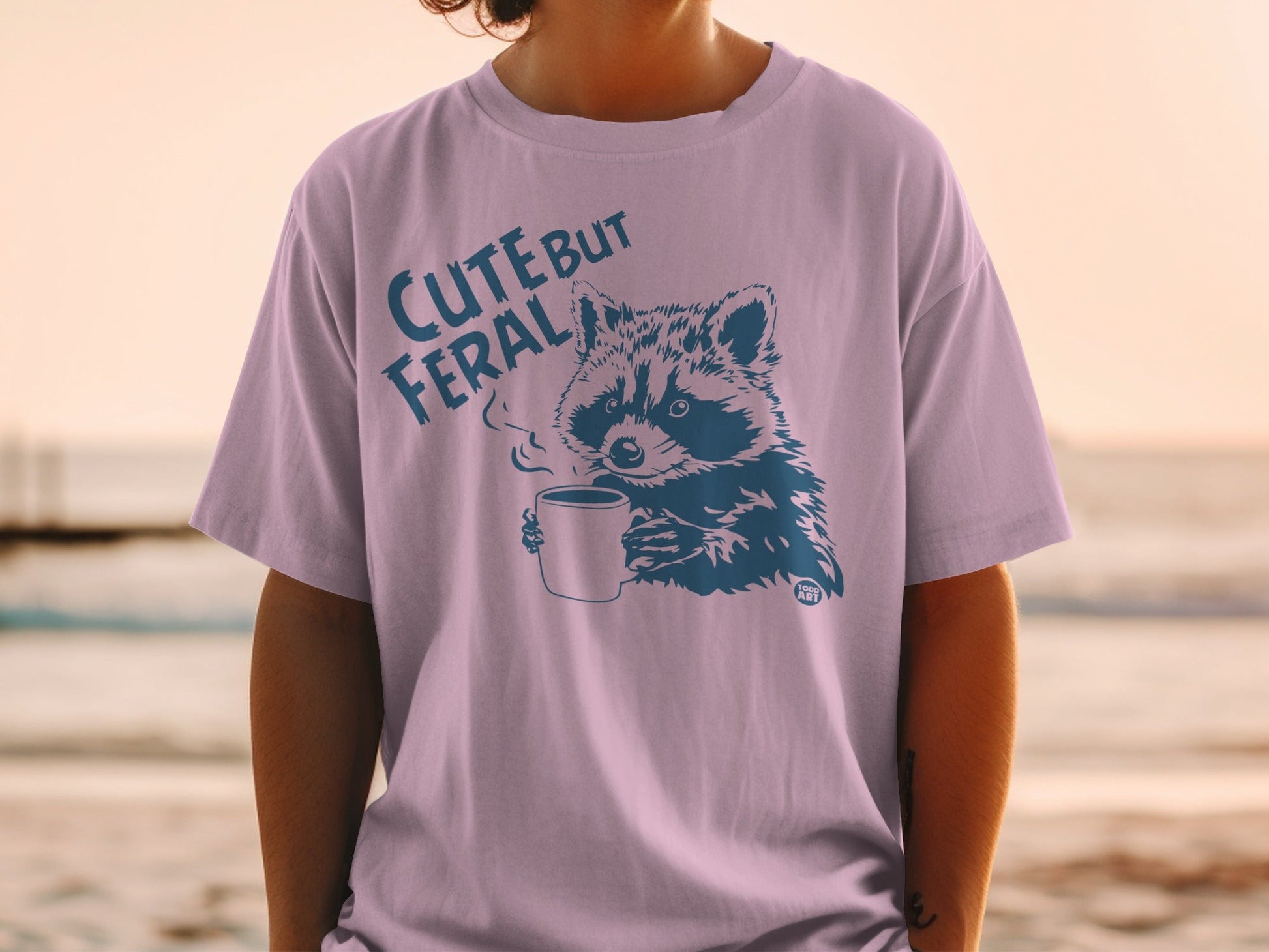 Blue raccoon with steam from coffee cup on light purple t-shirt.