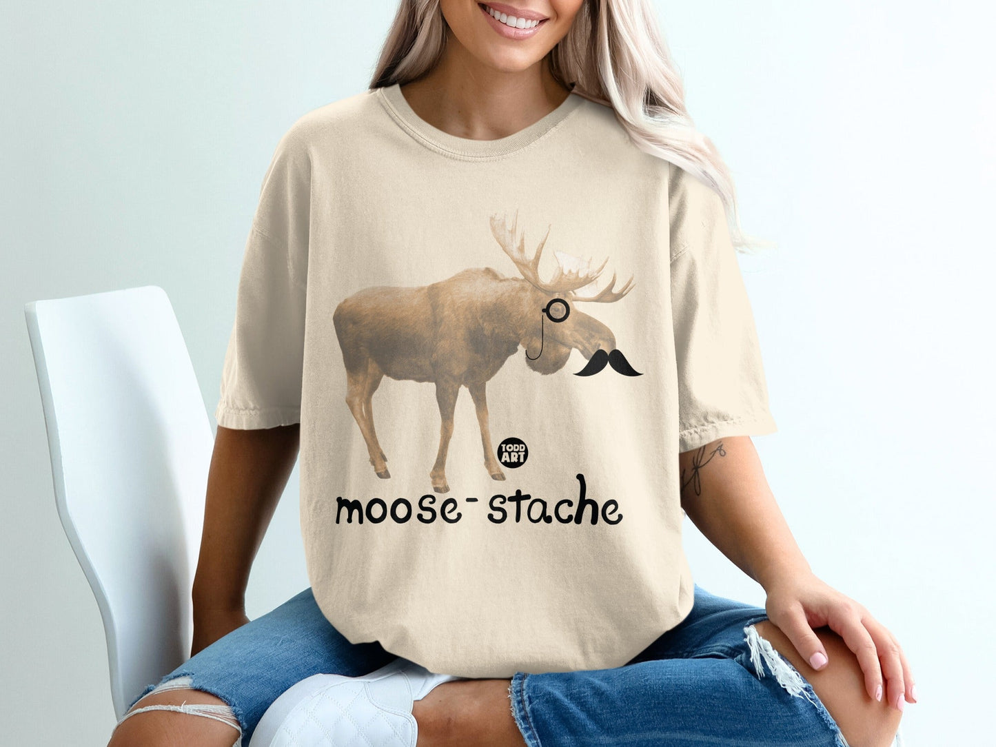 Moose with monocle and mustache on a cream t-shirt reading "moose-stache."