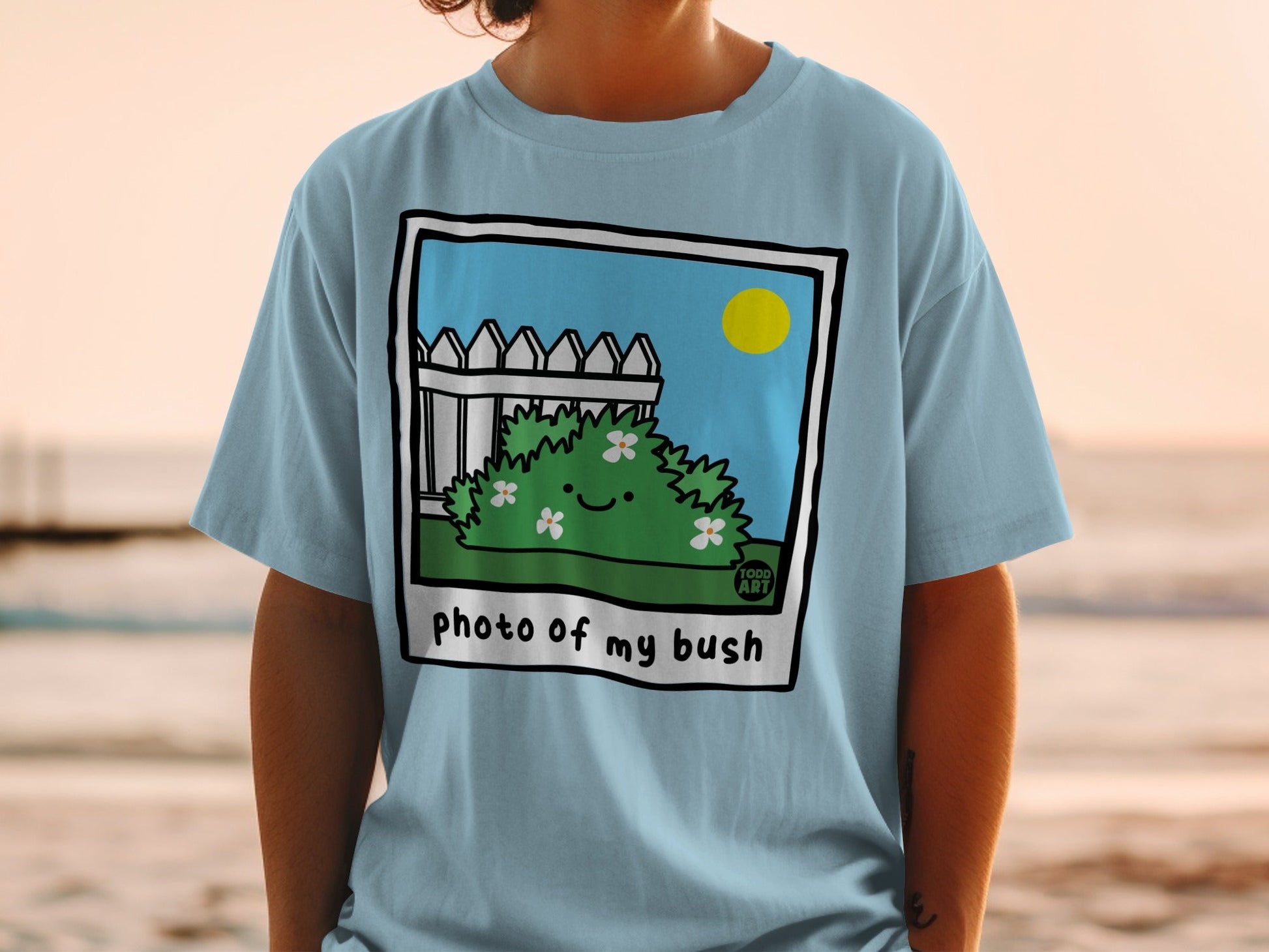 Light blue t-shirt with a cartoon bush and fence illustration reading "photo of my bush".