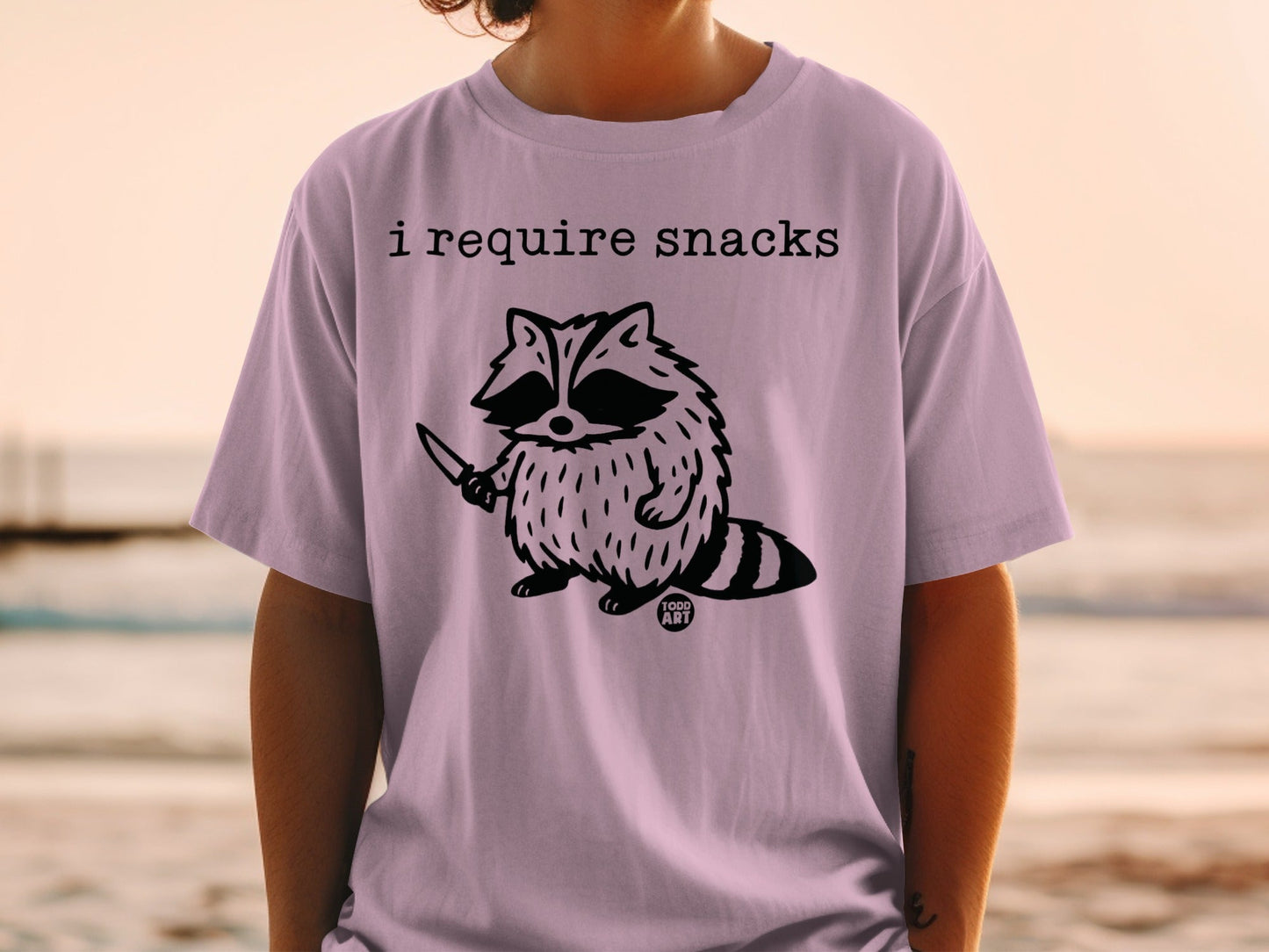 T-shirt with a raccoon holding a knife and the text "I require snacks".