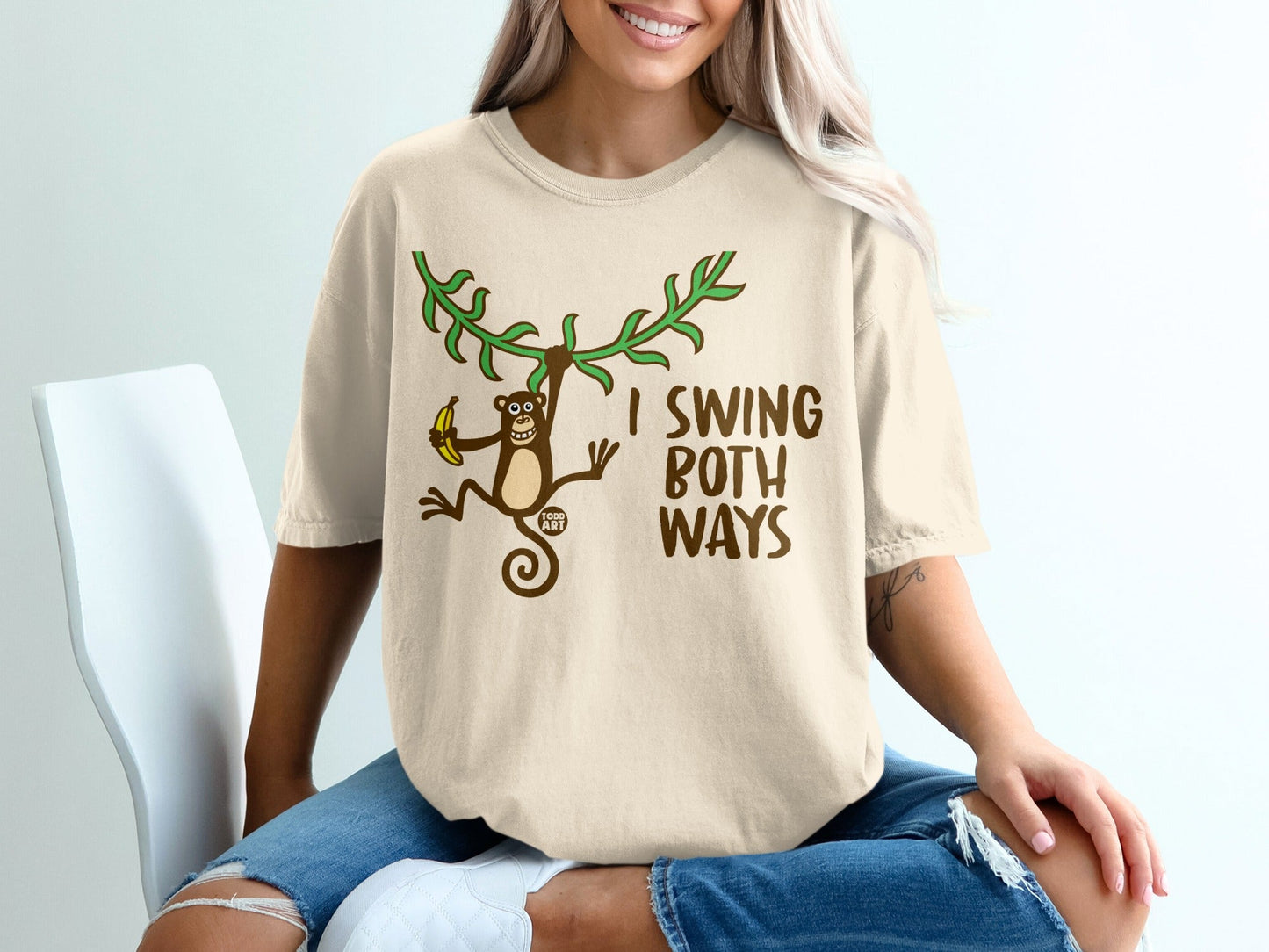 Beige t-shirt with a monkey hanging from branches holding a banana, text reads "I SWING BOTH WAYS".