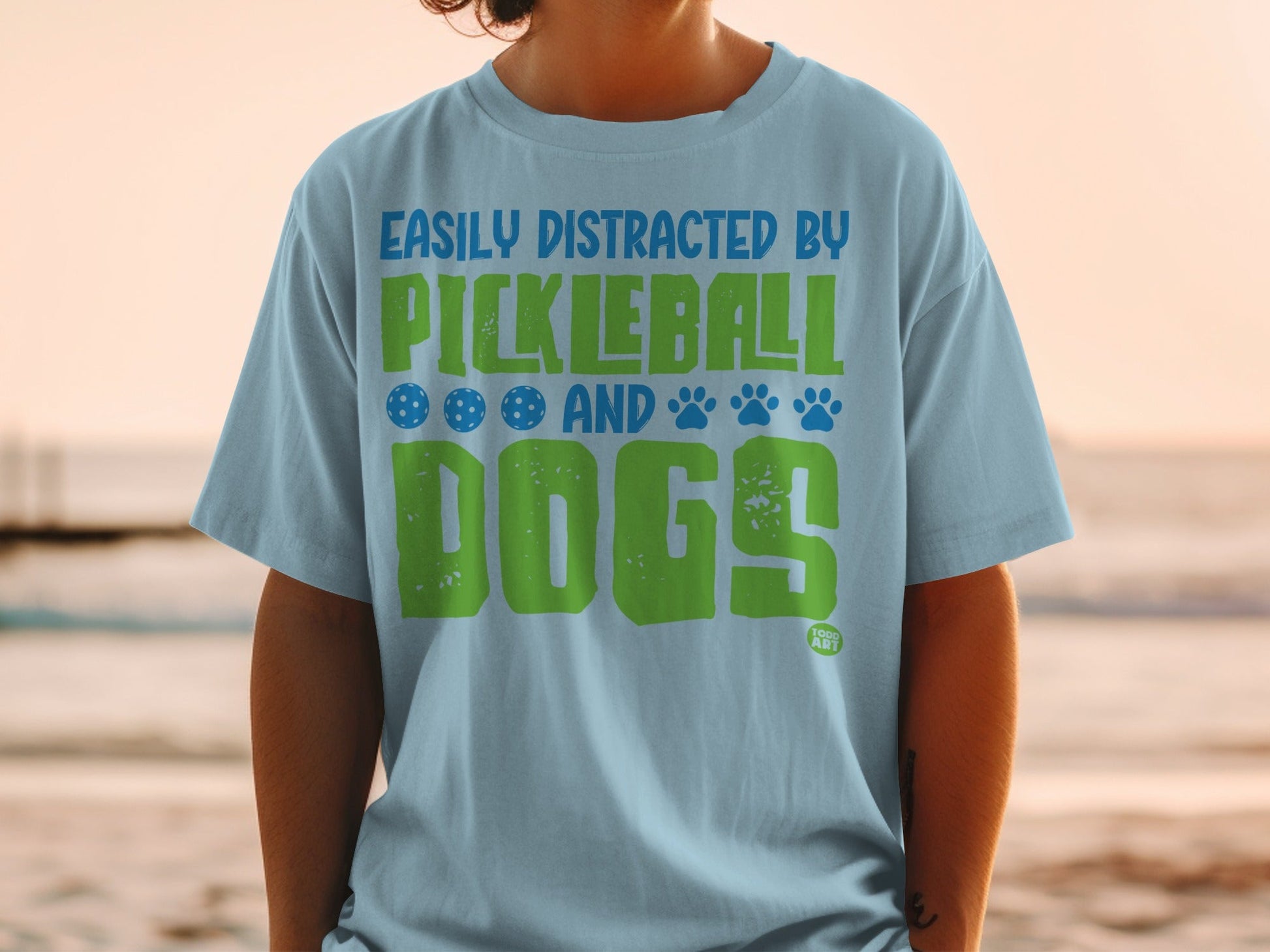 Light blue t-shirt with "Easily Distracted by Pickleball and Dogs" text and icons.
