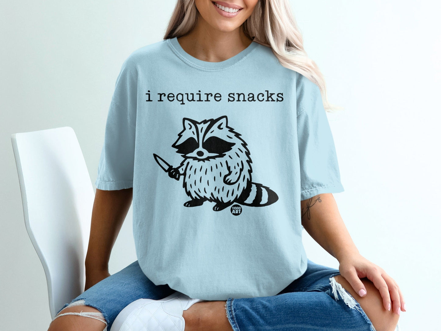 Light blue t-shirt with a raccoon holding a knife and text "i require snacks".