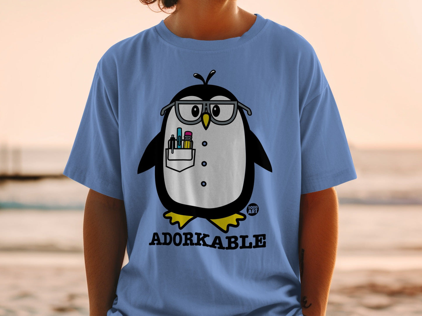 Light blue t-shirt with a cartoon penguin wearing glasses holding pens.