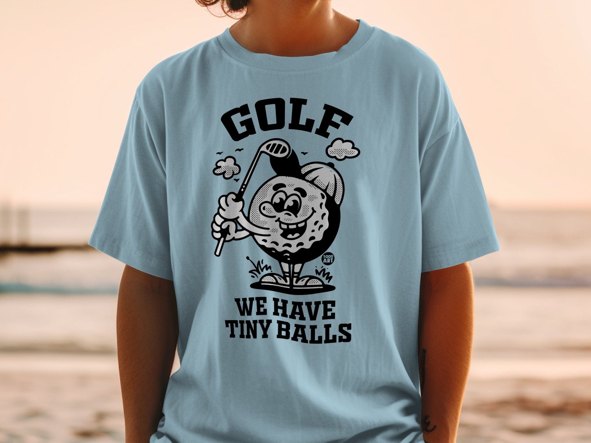 T-shirt with cartoon golf ball holding a club, reads "WE HAVE TINY BALLS".