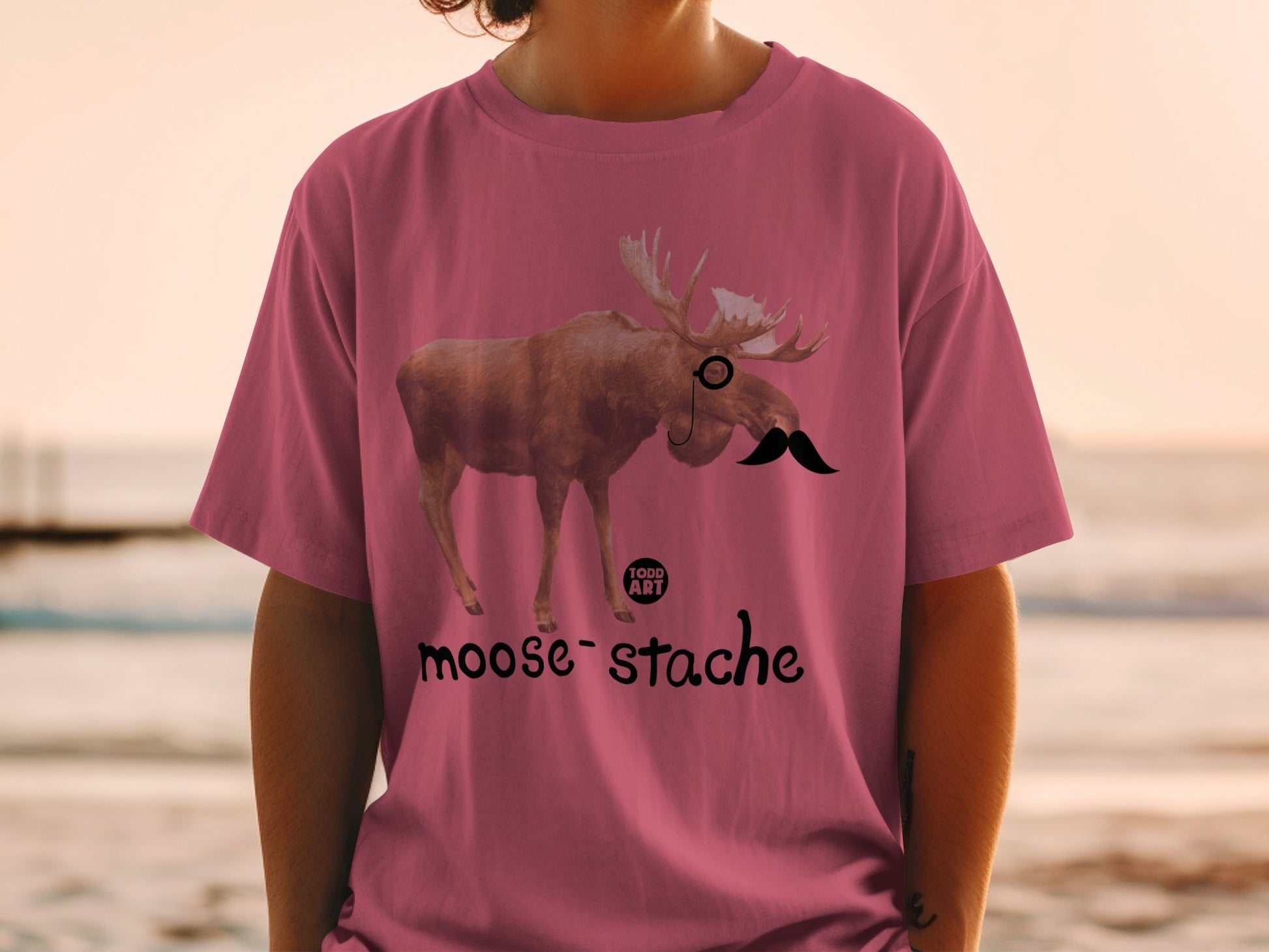Pink t-shirt with a moose wearing a monocle and mustache, "moose-stache" text.