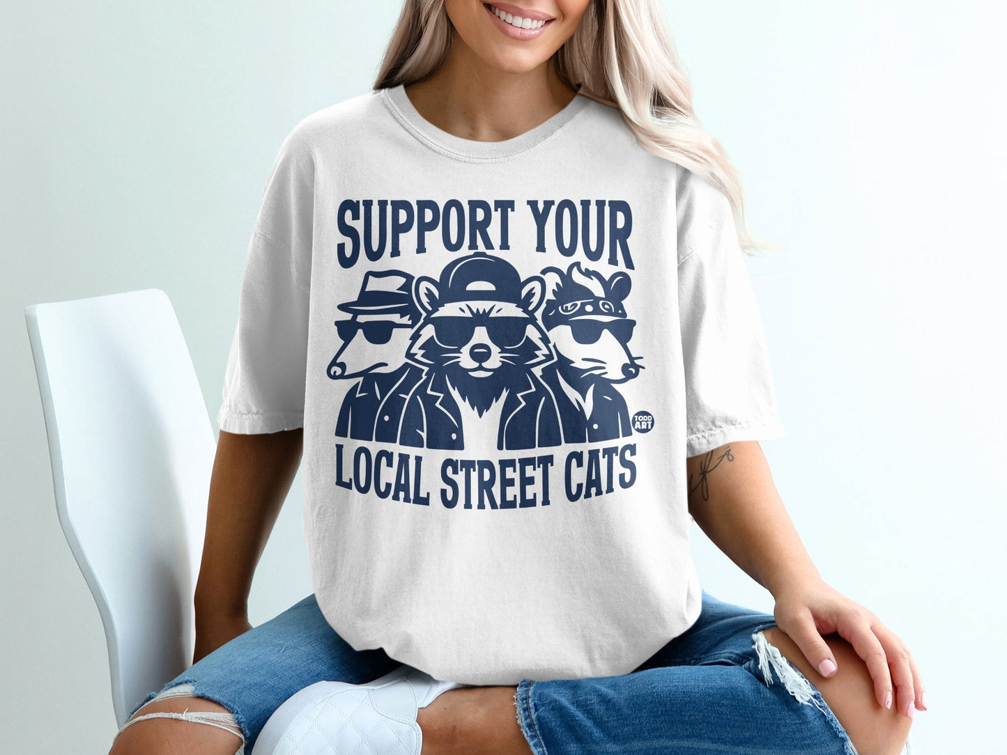 White t-shirt with navy graphic of three raccoons wearing sunglasses and hats, text "SUPPORT YOUR LOCAL STREET CATS".