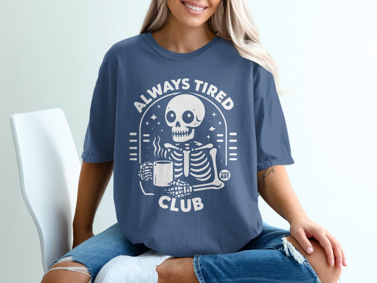 Skeleton holding coffee mug with "Always Tired Club" text on navy t-shirt.