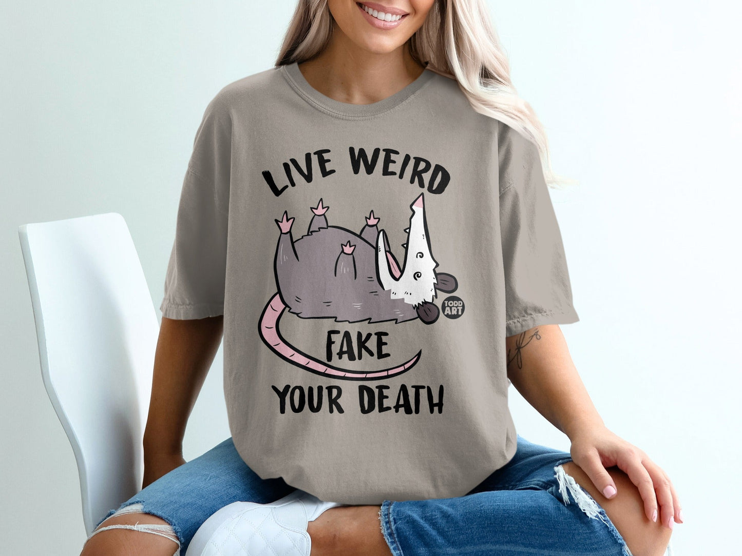 T-shirt with a gray opossum playing dead, text "LIVE WEIRD FAKE YOUR DEATH".