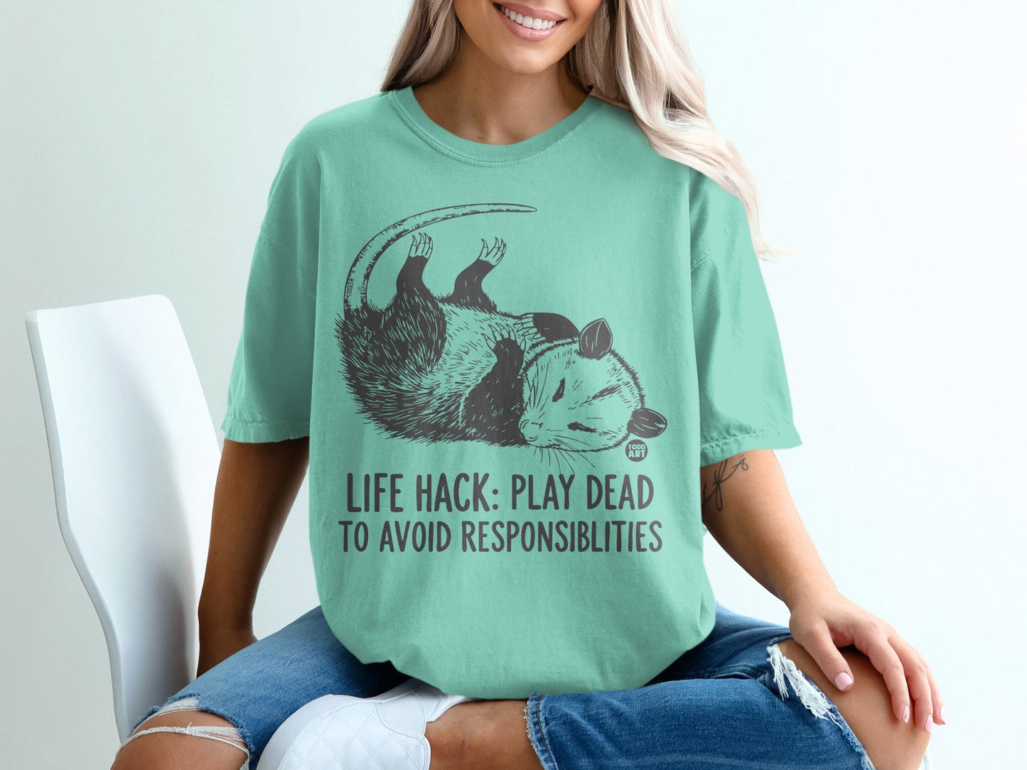 Opossum with "LIFE HACK: PLAY DEAD TO AVOID RESPONSIBILITIES" text on teal t-shirt.