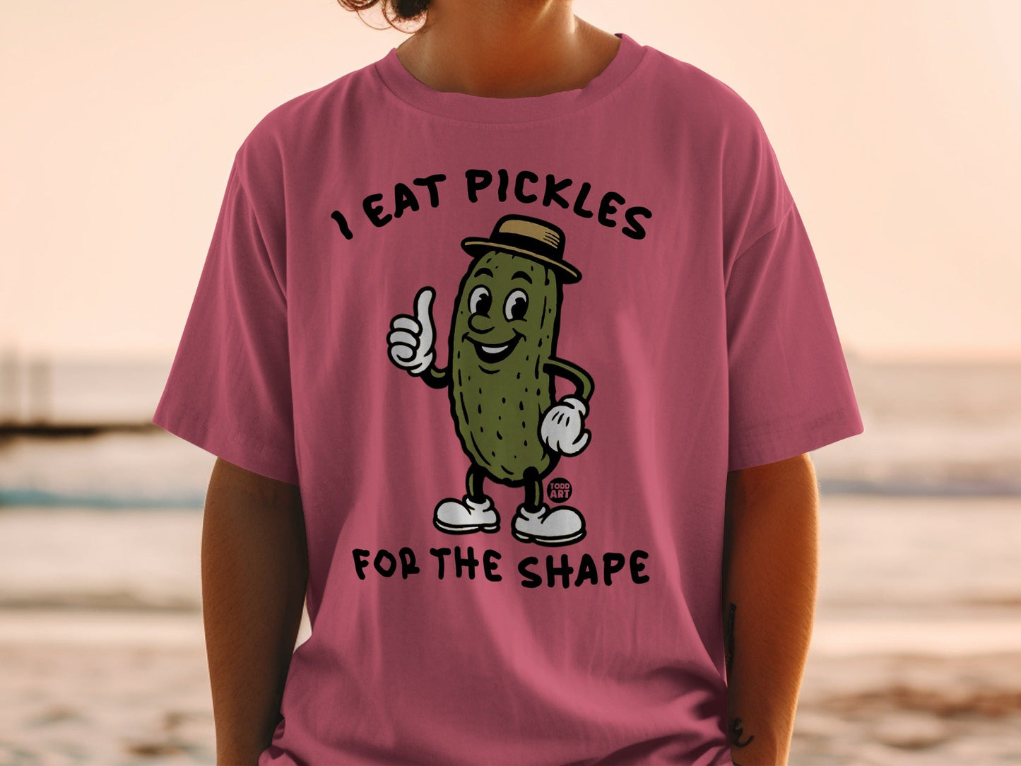 Cartoon pickle wearing a hat gives a thumbs up on a pink t-shirt.