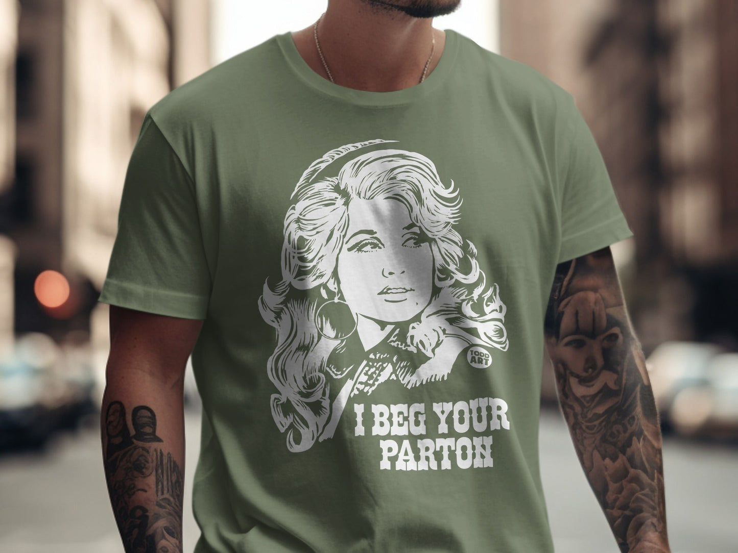Olive green t-shirt featuring a black and white illustration of Dolly Parton with text "I BEG YOUR PARTON".