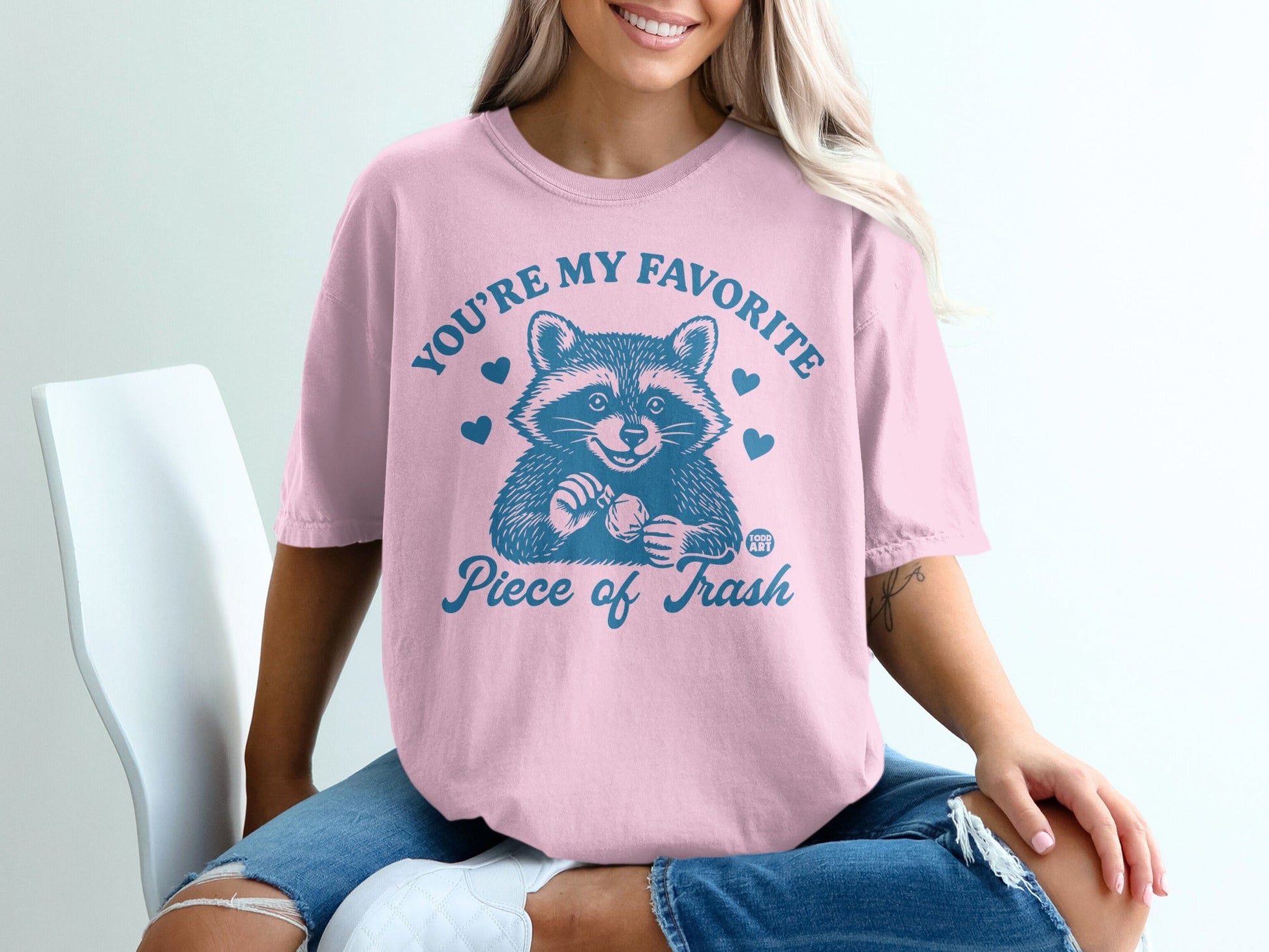 Pink t-shirt with raccoon illustration and text "You're my favorite piece of trash."