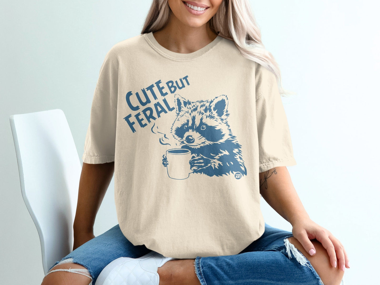 Cream t-shirt with a raccoon drinking coffee illustration and "Cute but Feral" text.