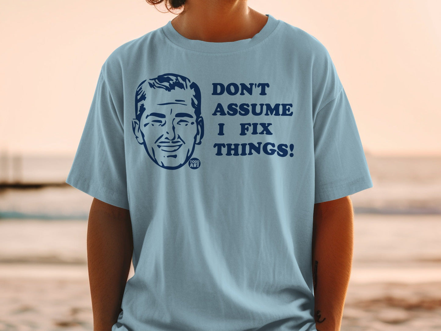 Light blue t-shirt with a cartoon man's face and text "DON'T ASSUME I FIX THINGS!".