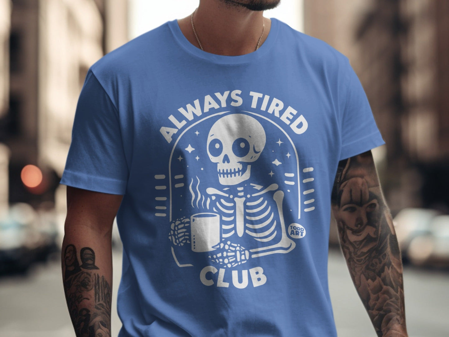 Blue t-shirt with a skeleton holding a coffee mug and "Always Tired Club" text.