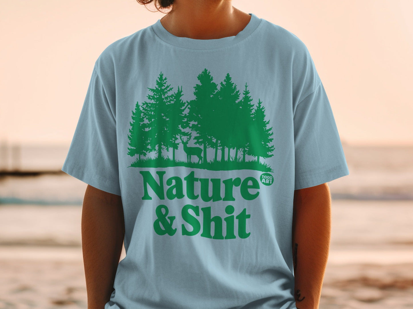 Light blue t-shirt with green forest and deer graphic, text "Nature & Shit".
