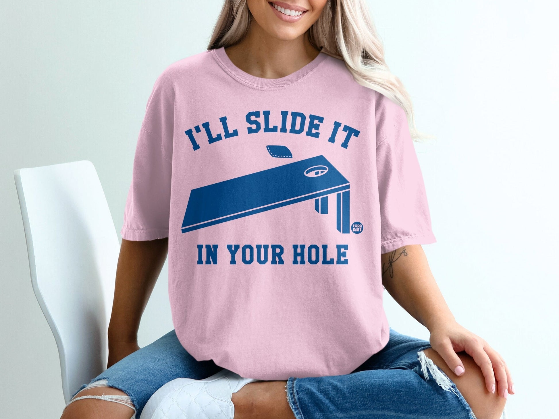 Pink t-shirt with blue text "I'LL SLIDE IT IN YOUR HOLE" and a cornhole board.
