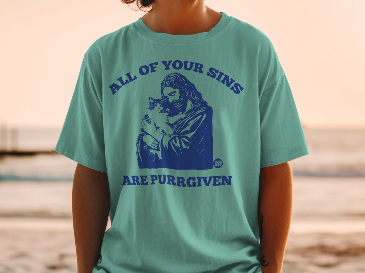 Blue graphic t-shirt with Jesus holding a cat and text "ALL OF YOUR SINS ARE PURRGIVEN".
