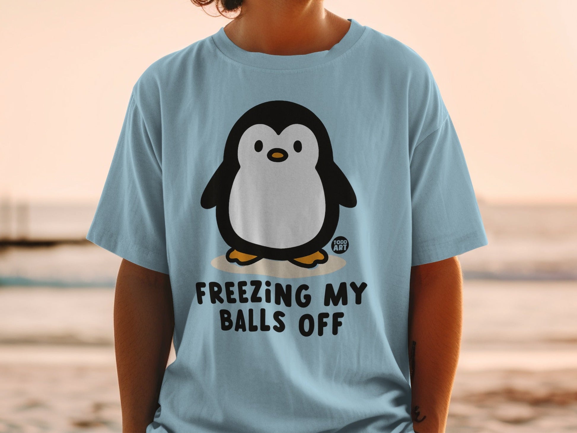 Light blue t-shirt with a cartoon penguin and text "FREEZING MY BALLS OFF".