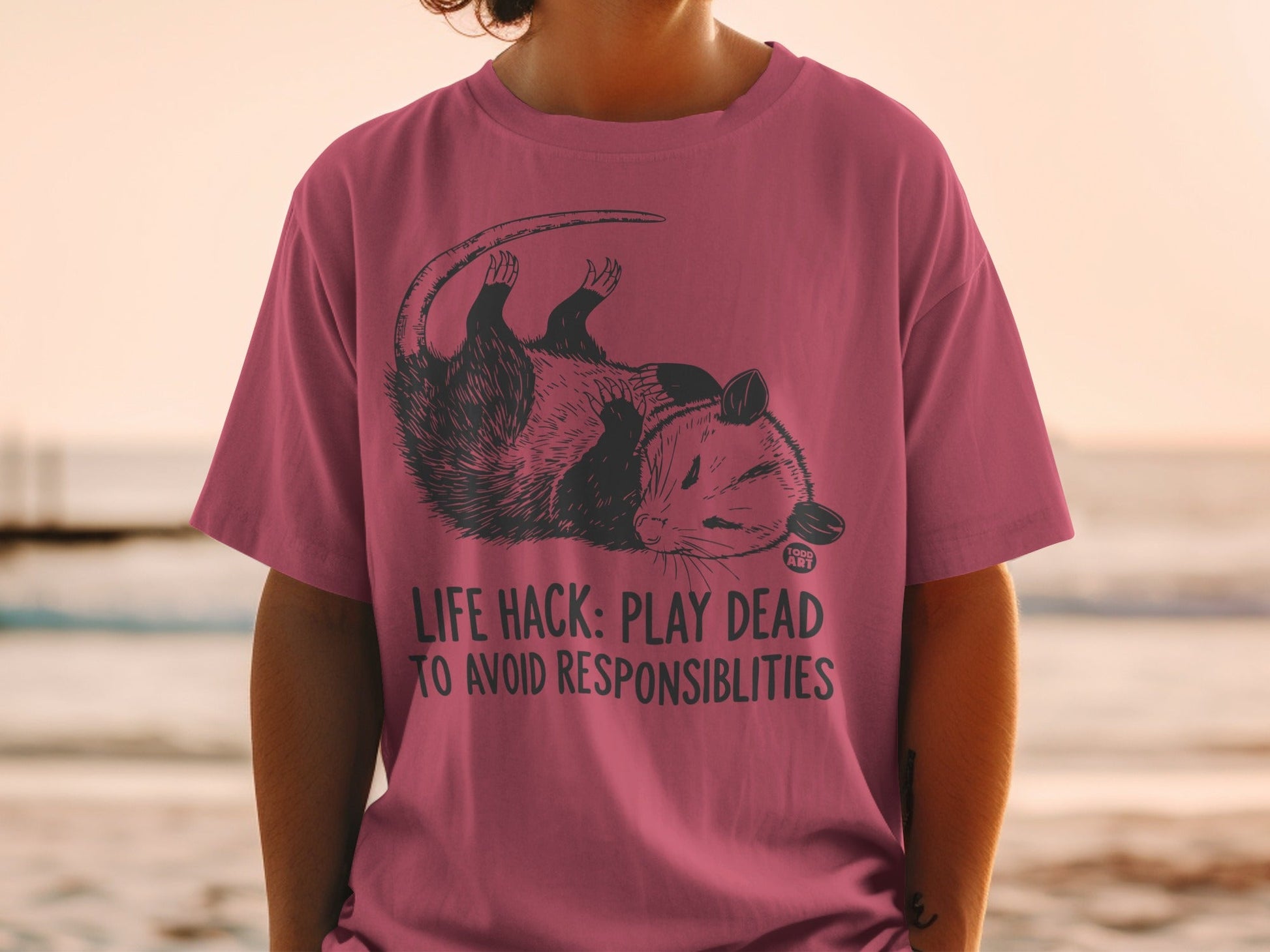 Pink t-shirt with a possum and text "Life Hack: Play Dead to Avoid Responsibilities".
