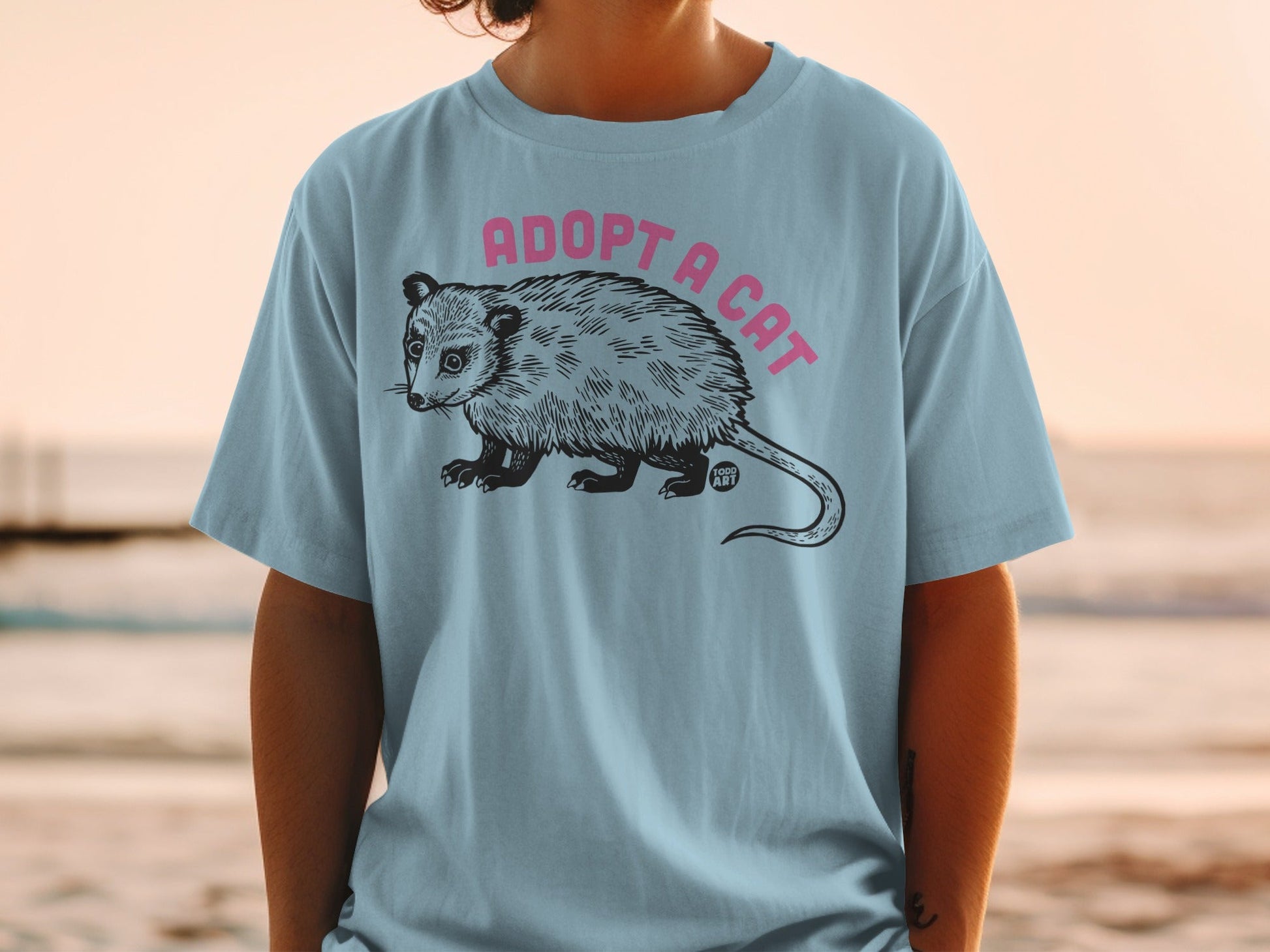 Light blue t-shirt featuring a black and white illustration of an opossum with "ADOPT A CAT" in pink text above.