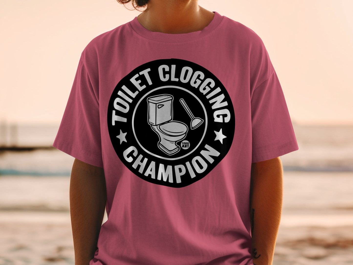 Circular graphic on pink t-shirt with toilet, plunger, and "Toilet Clogging Champion" text.