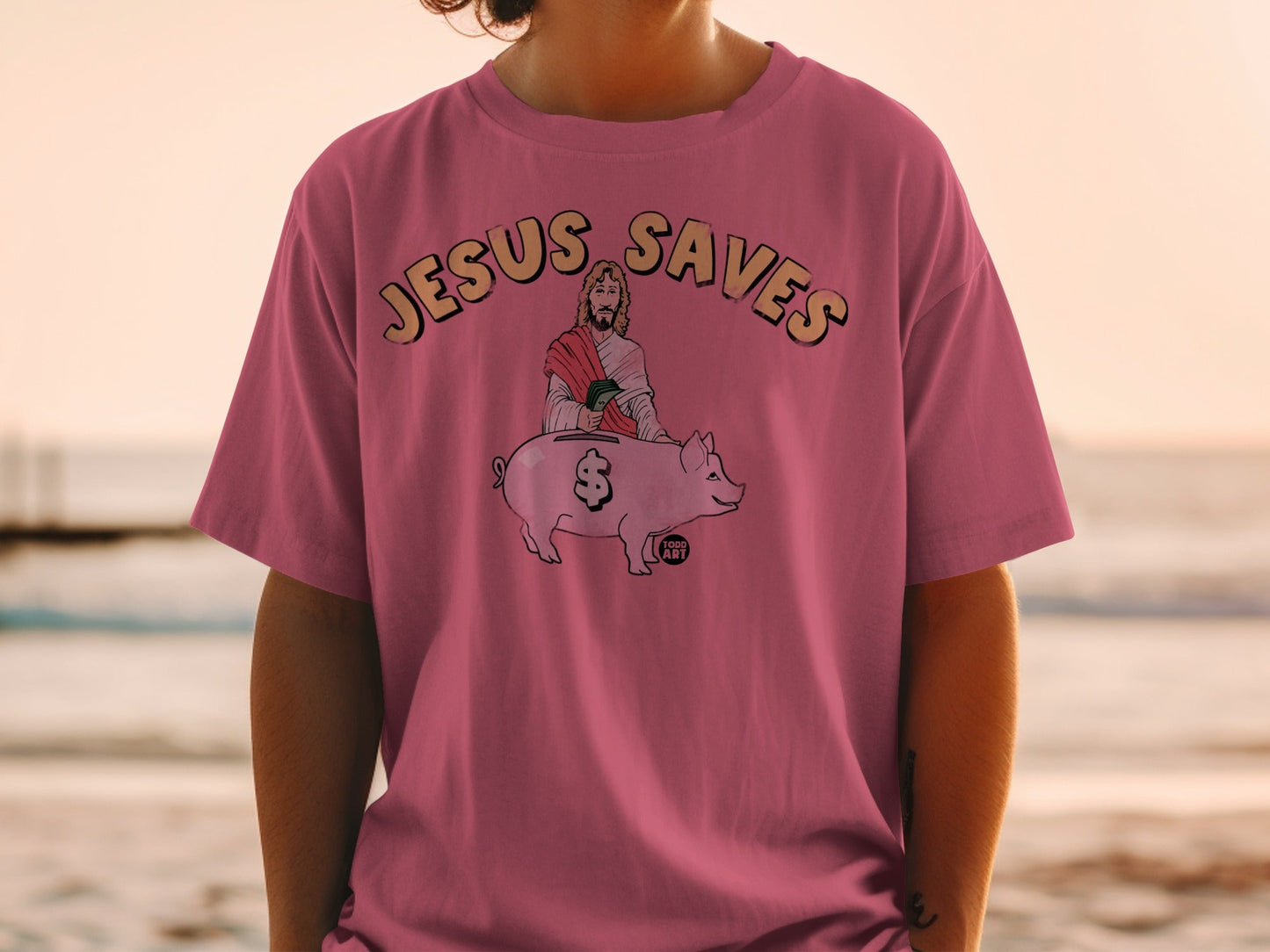 Pink t-shirt with "Jesus Saves" text and illustration of Jesus hugging a piggy bank.