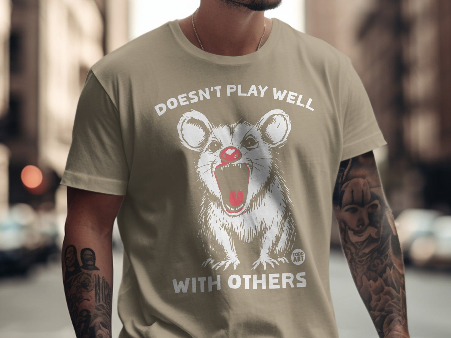 Tan t-shirt with white graphic of an opossum with red nose and text "DOESN'T PLAY WELL WITH OTHERS".