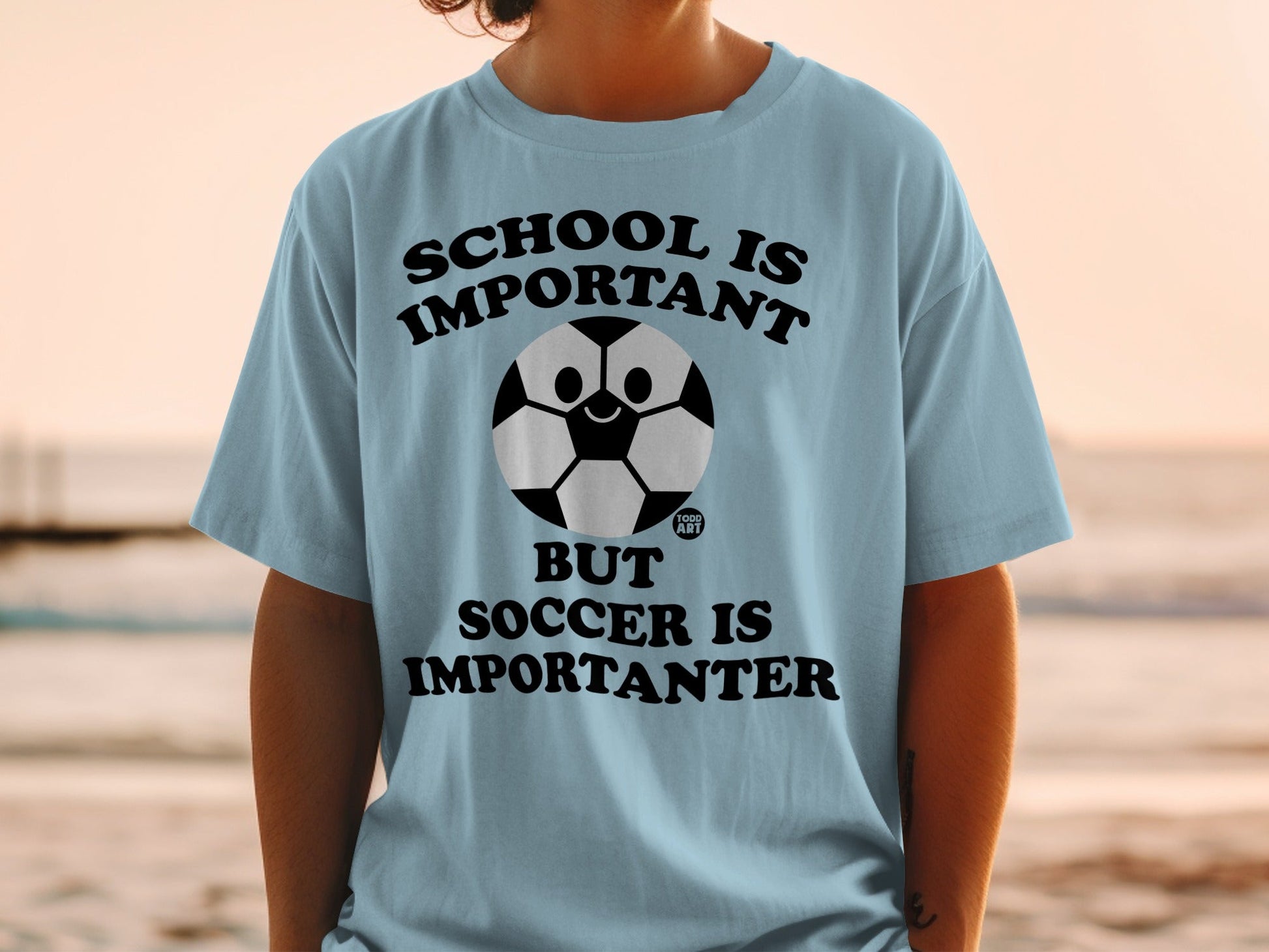 Light blue t-shirt with text "SCHOOL IS IMPORTANT BUT SOCCER IS IMPORTANTER" and a smiling soccer ball.