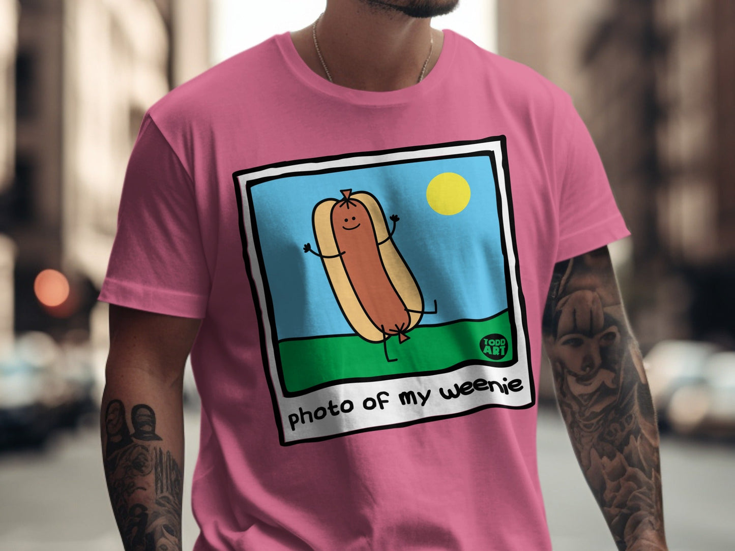 Pink t-shirt featuring a cartoon hot dog in a sunny outdoor scene.