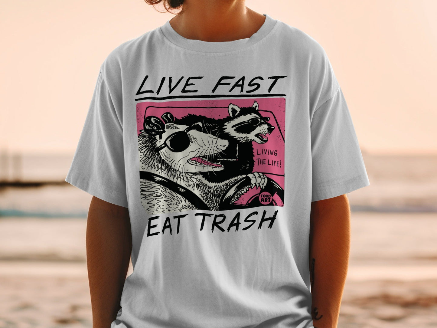 Graphic tee with raccoon and possum driving, "LIVE FAST EAT TRASH" text.