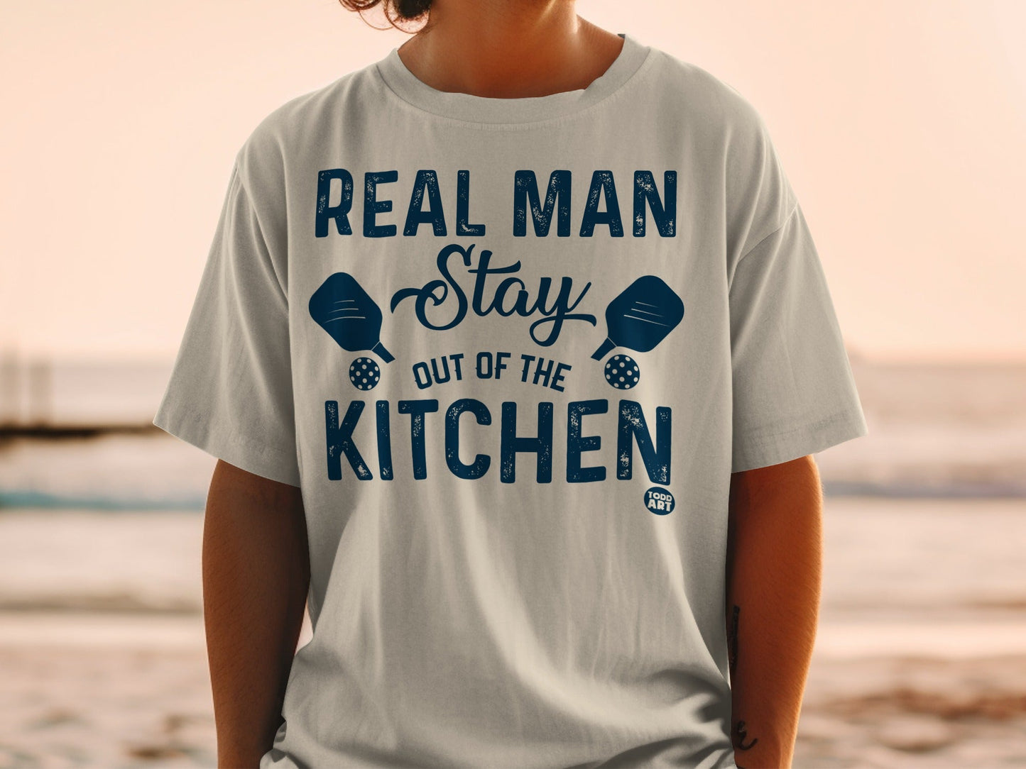 T-shirt with text "REAL MAN Stay OUT OF THE KITCHEN" and pickleball paddles.