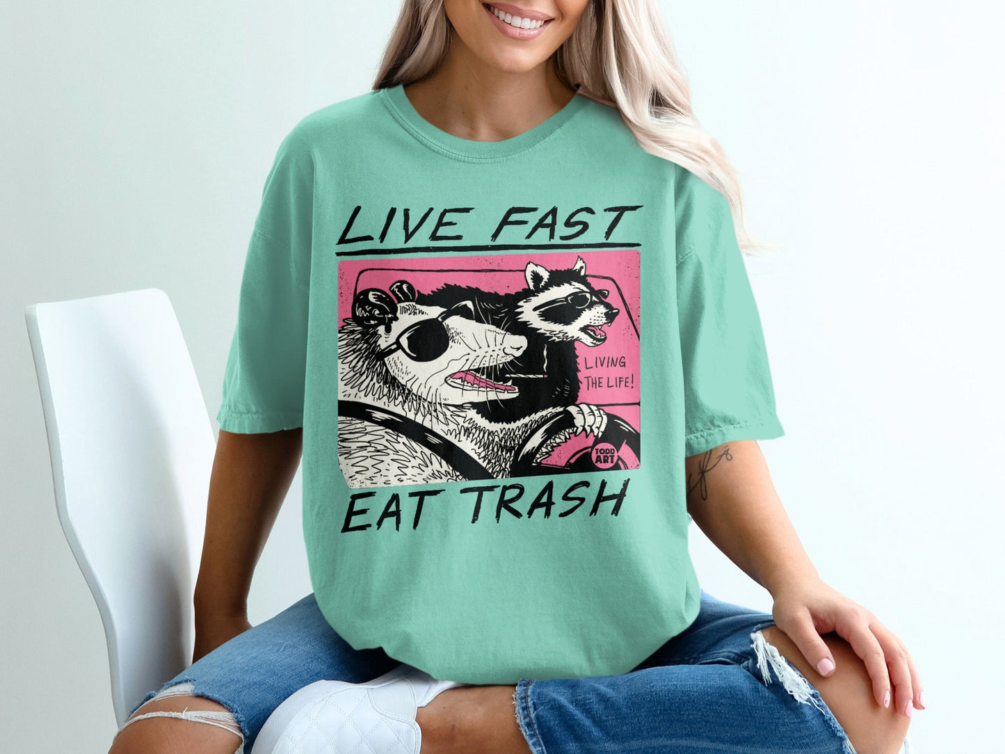 Mint green t-shirt with "Live Fast Eat Trash" and cartoon raccoons driving.