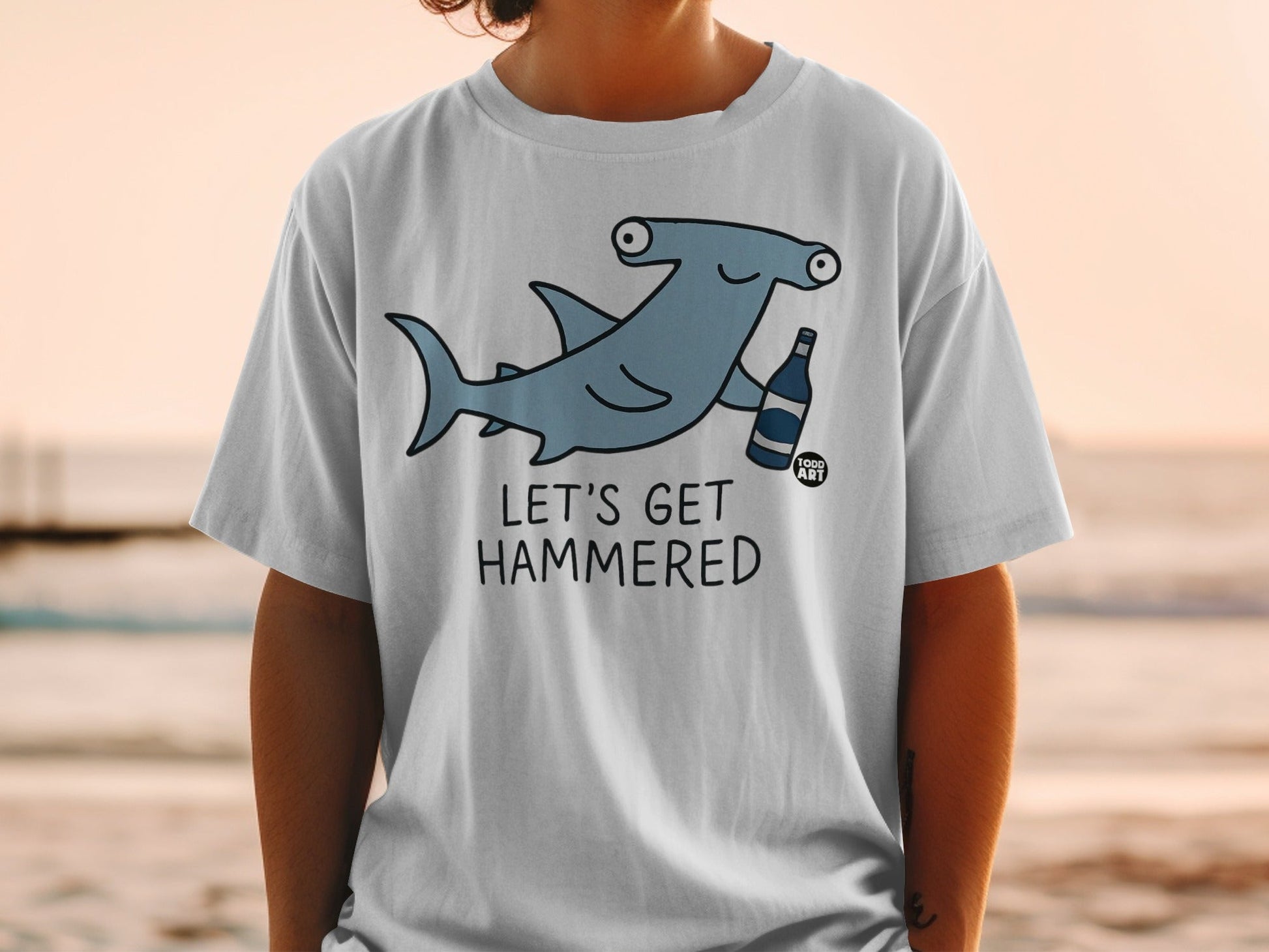 T-shirt graphic of a cartoon hammerhead shark holding a bottle, with text "LET'S GET HAMMERED".