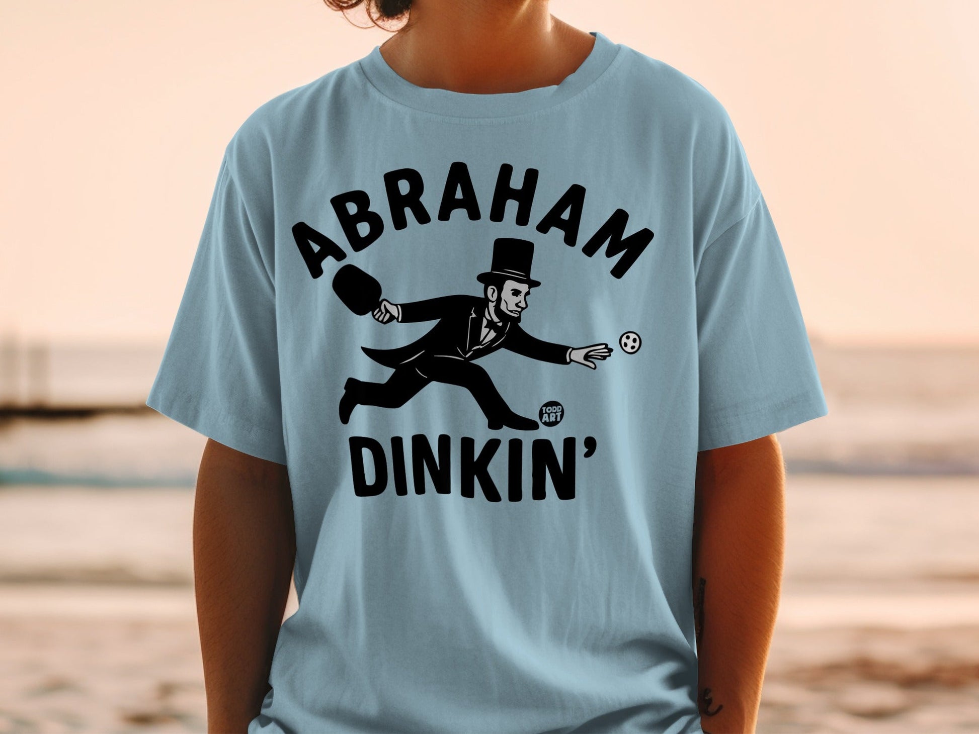 T-shirt with Abraham Lincoln playing pickleball, text "ABRAHAM DINKIN'".