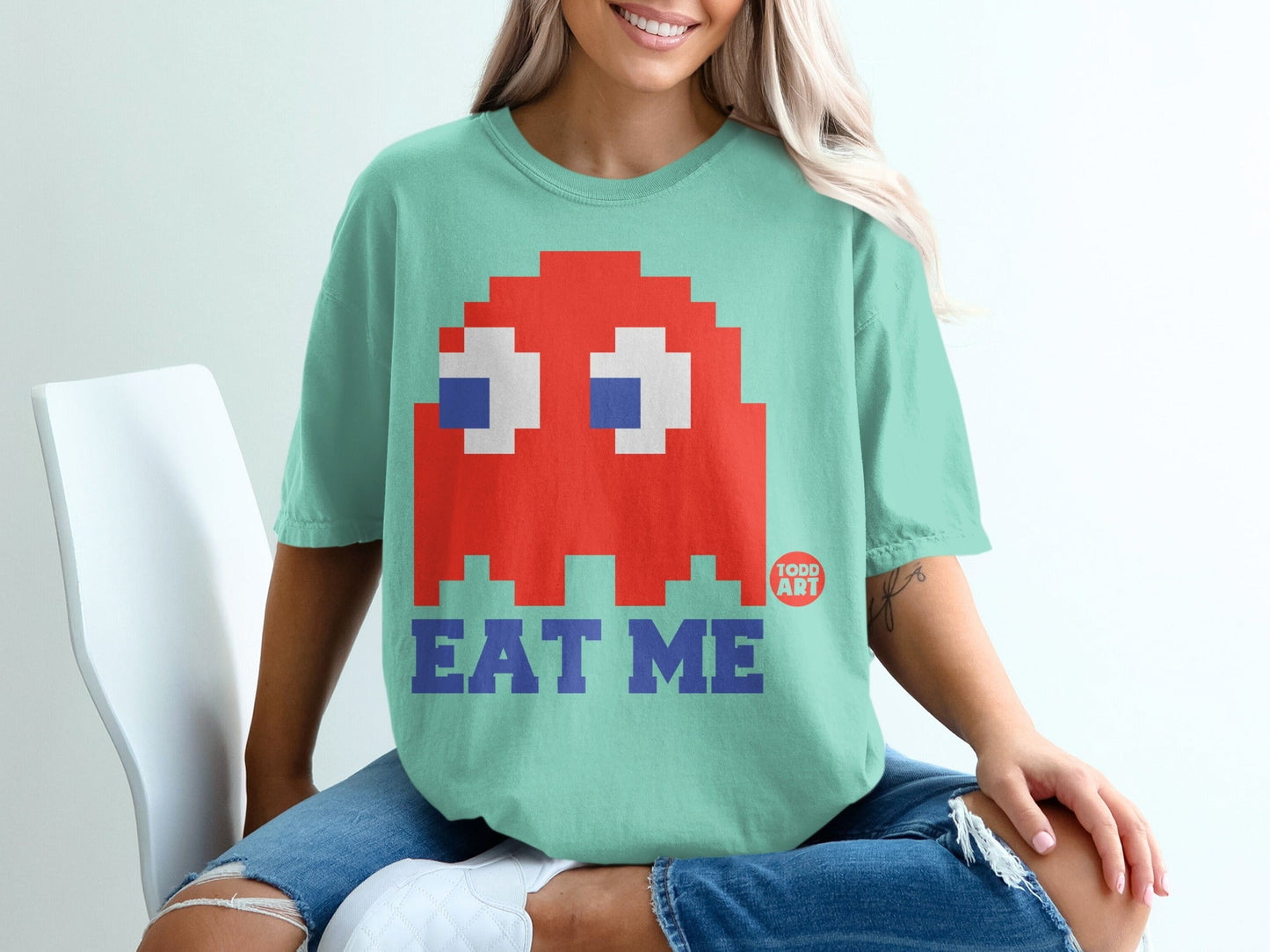 Red pixelated Pac-Man ghost graphic on mint green t-shirt with "EAT ME" text.