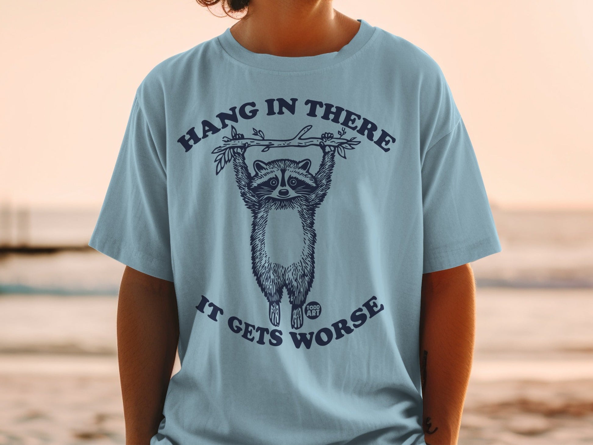 T-shirt with raccoon hanging from branch, text "Hang in there it gets worse".