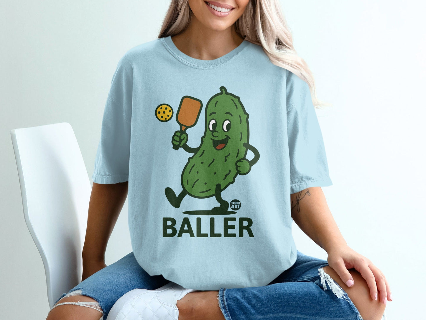 Light blue t-shirt featuring cartoon pickle holding paddle and ball, text "BALLER".