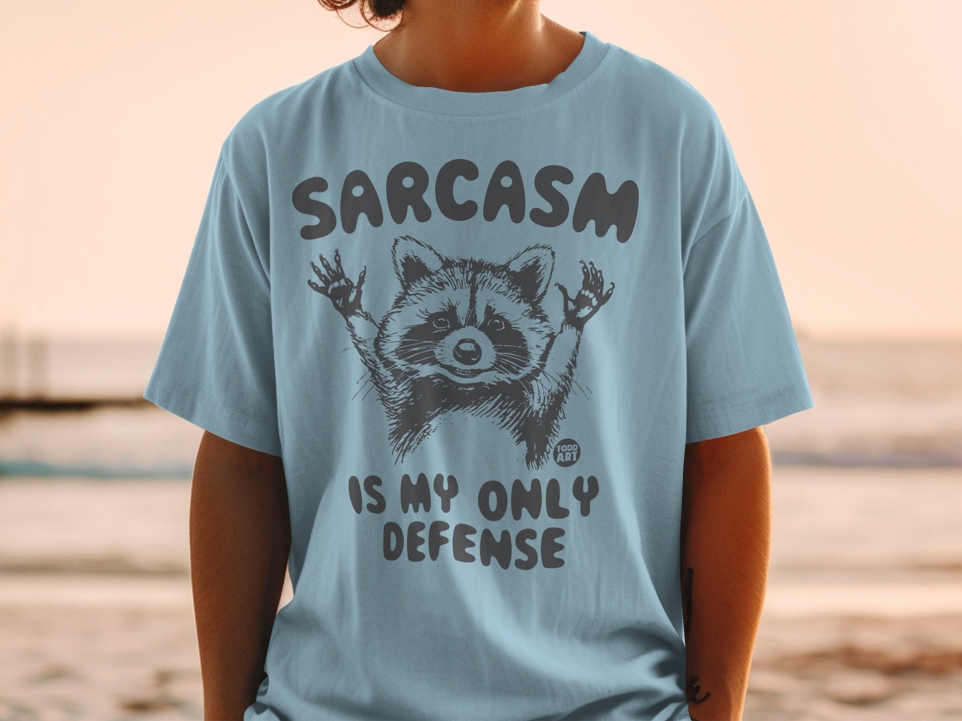 Light blue t-shirt with raccoon graphic and text "SARCASM IS MY ONLY DEFENSE".