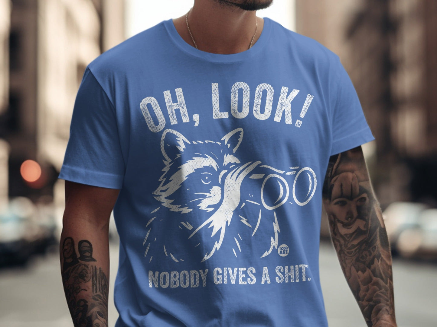 Blue t-shirt with white raccoon looking through binoculars and text "OH, LOOK! NOBODY GIVES A SHIT."
