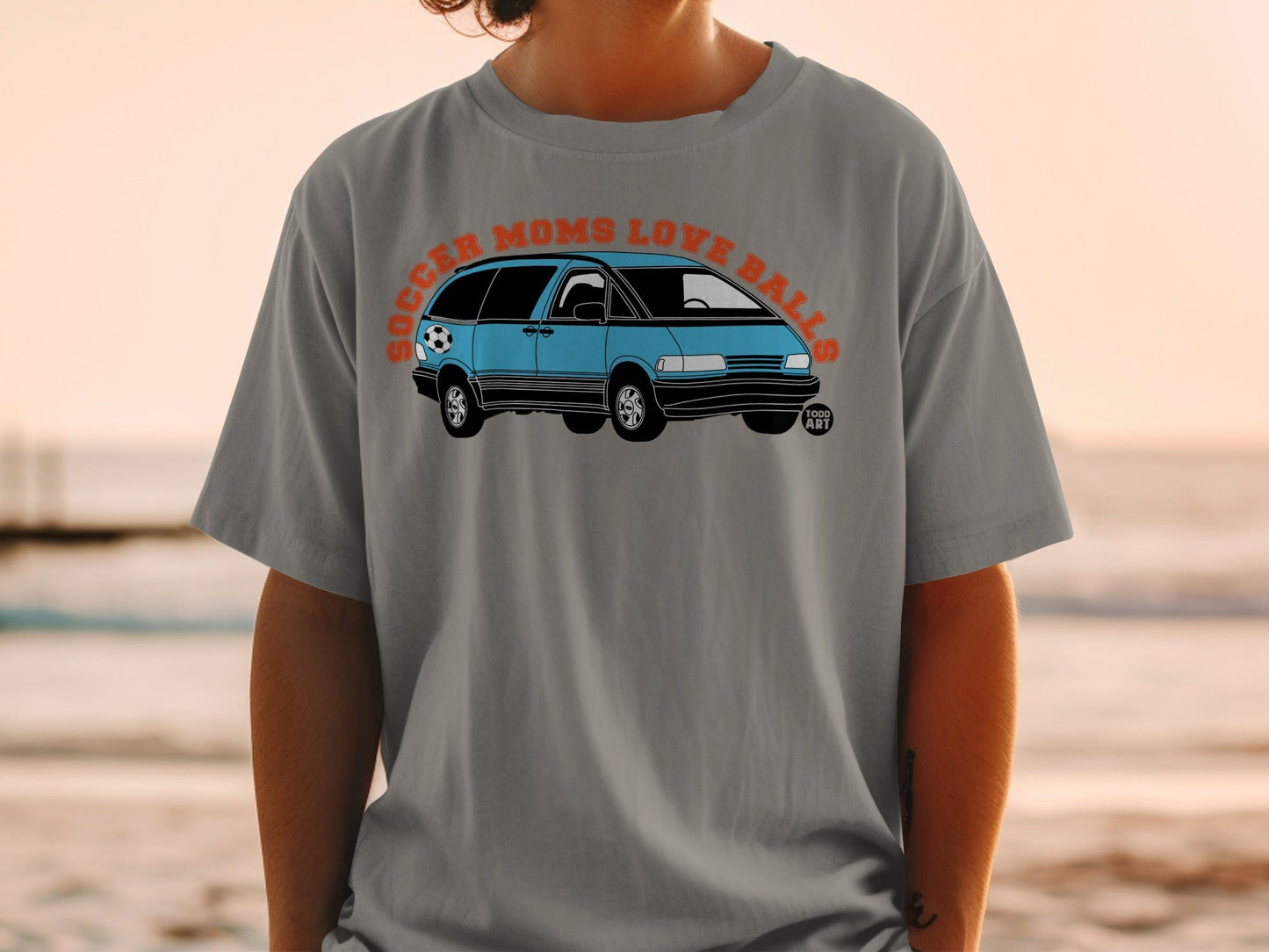 Gray t-shirt with a blue minivan and soccer ball graphic, text reads "SOCCER MOMS LOVE BALLS".