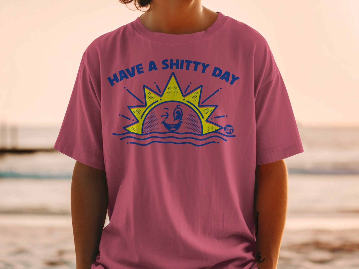 Pink t-shirt with blue text "HAVE A SHITTY DAY" and winking sun graphic.